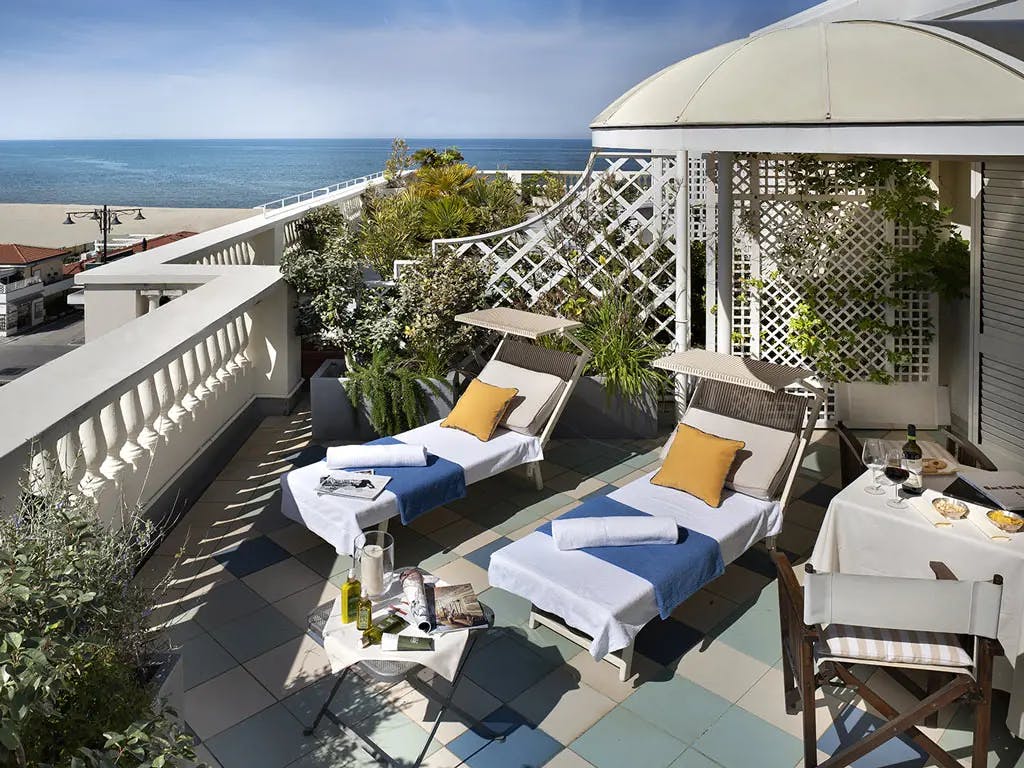 A private terrace features two lounge chairs with blue and yellow accents, surrounded by greenery. A dining table with two chairs is set with snacks, while a scenic lateral view of the sea is visible in the background under a clear sky.