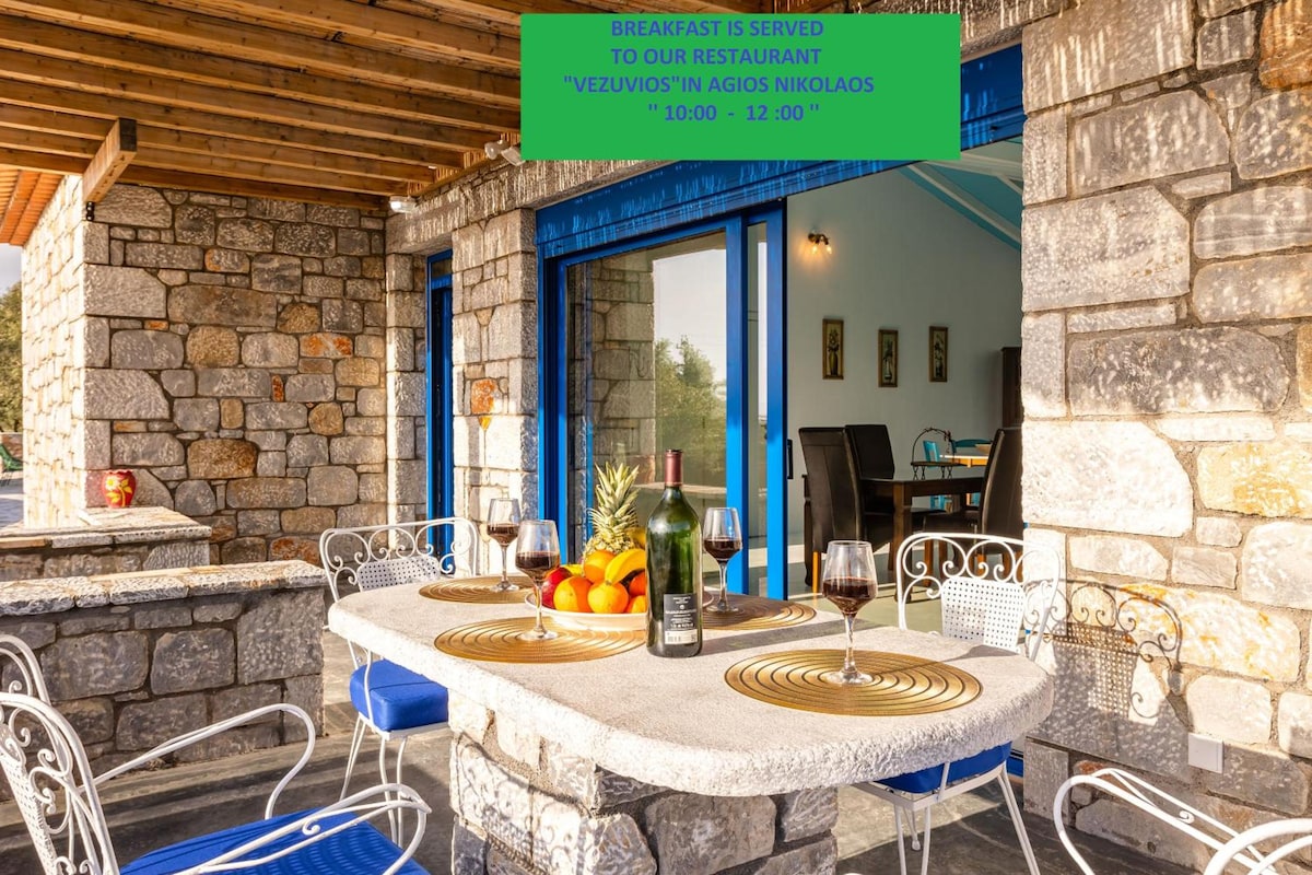 An outdoor dining area features a stone table set with wine glasses and a bottle, surrounded by fresh fruit. Bright blue doors open to reveal a view of the natural surroundings, contrasting with the rustic stone walls and wooden beams overhead.