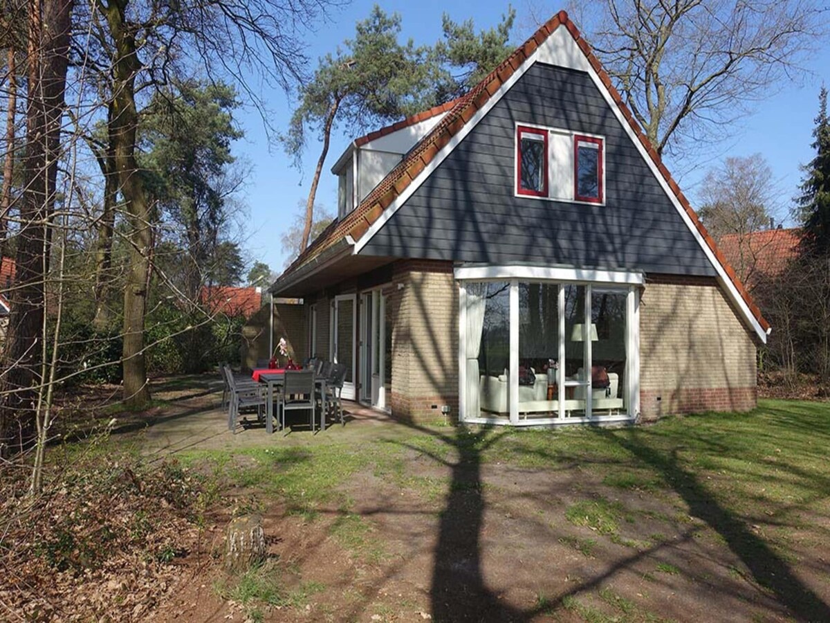 A modern two-story house is surrounded by trees, featuring a large outdoor patio area with a dining table and chairs. Expansive glass doors provide a view into the interior space, which is illuminated by natural light. The exterior boasts a sloped roof with red accents.