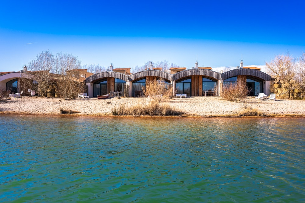 A row of holiday houses is positioned along a sandy beach, offering direct access to tranquil waters. Each house features large windows and a wooden facade, surrounded by sparse vegetation. The calm lake reflects a clear blue sky, enhancing the serene atmosphere.