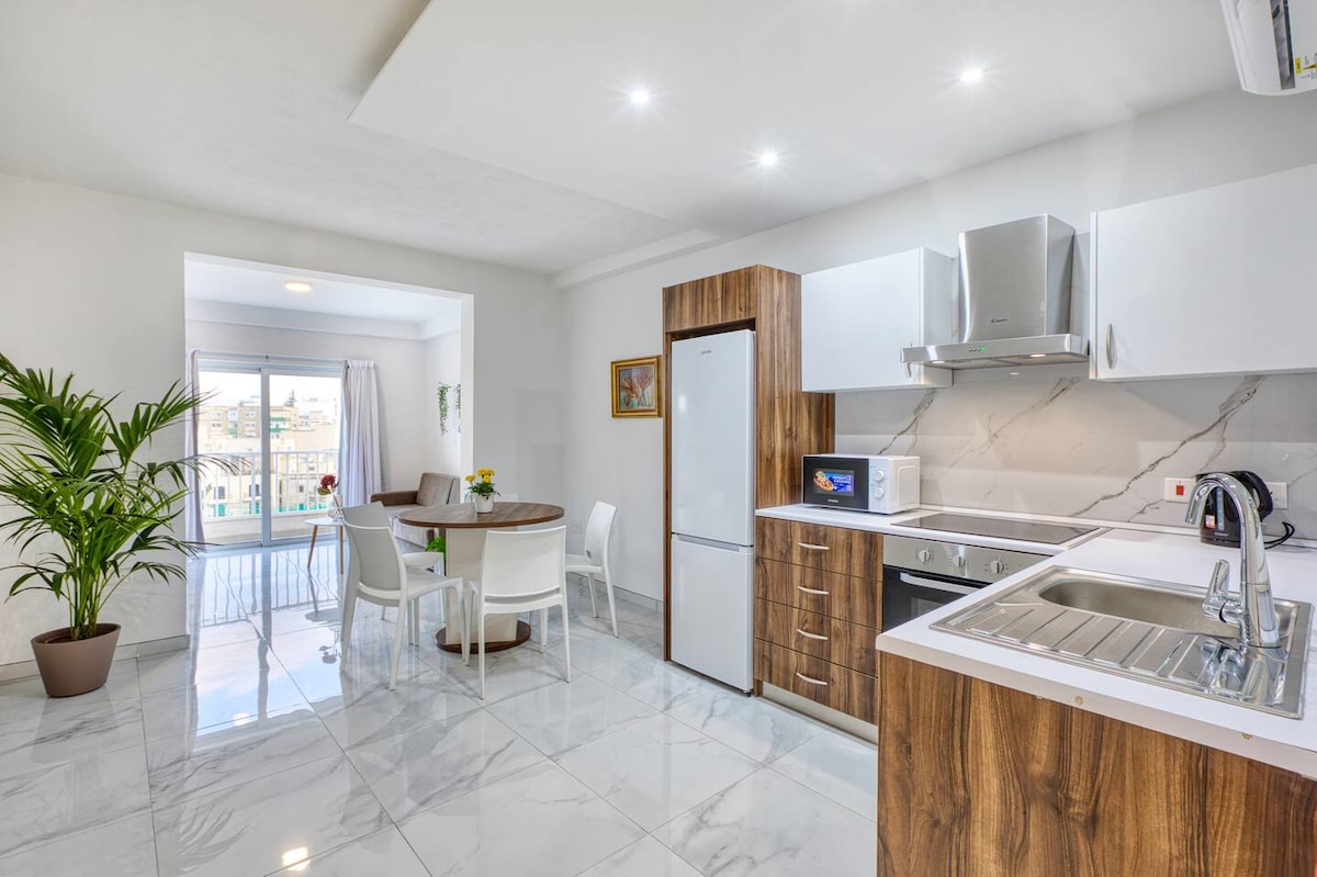 A modern kitchen area features sleek wooden cabinetry and a marble countertop. A round dining table is surrounded by four white chairs. Large windows allow natural light to fill the space, offering views of the balcony and greenery outside. A potted plant adds a touch of nature.