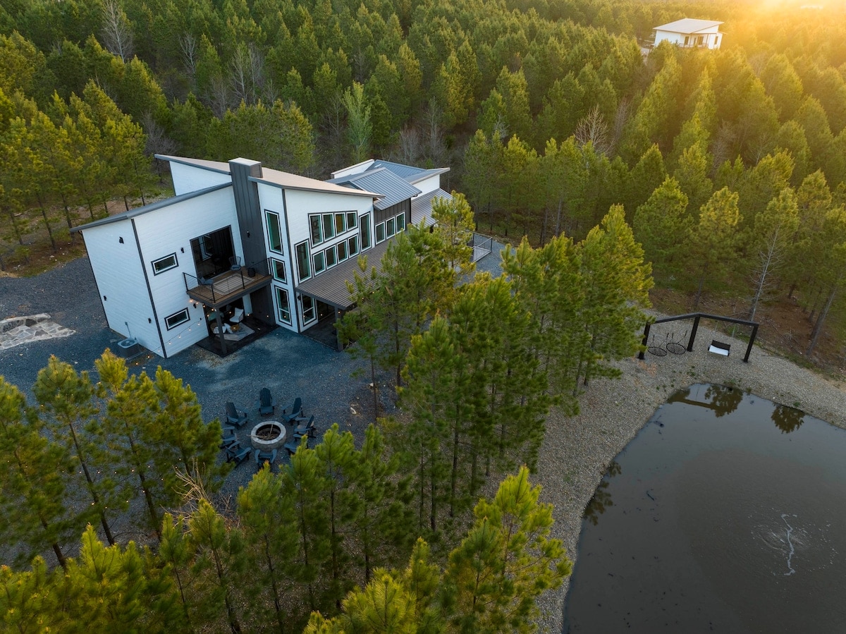 An aerial view captures a modern cabin surrounded by dense evergreen trees and a tranquil pond. A circular fire pit area is visible near the water's edge, complemented by the setting sun casting a warm glow over the landscape.