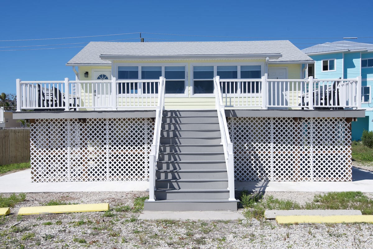 A charming upper duplex features a light yellow exterior with white railings and a spacious front deck. Stairs lead up to the entrance, framed by decorative lattice work below. Clear blue skies provide a bright backdrop to this inviting space.