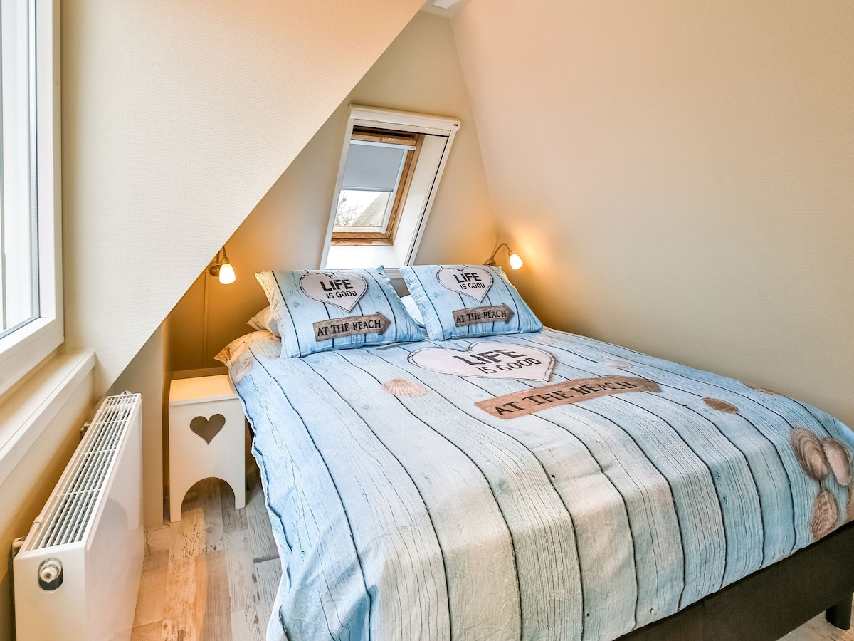 A cozy bedroom features a slanted ceiling and a spacious bed adorned with beach-themed bedding. Soft lighting is provided by wall-mounted lamps on either side of the bed. A small bedside table with a heart-shaped cutout adds charm, while a window allows natural light to fill the space.