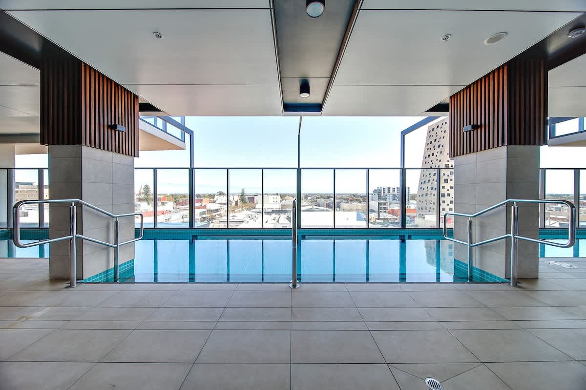 A heated swimming pool is situated within a modern indoor space, framed by large glass windows offering expansive views of the surrounding area. The pool is surrounded by sleek tiled flooring and comfortable access rails, creating a serene environment for relaxation.