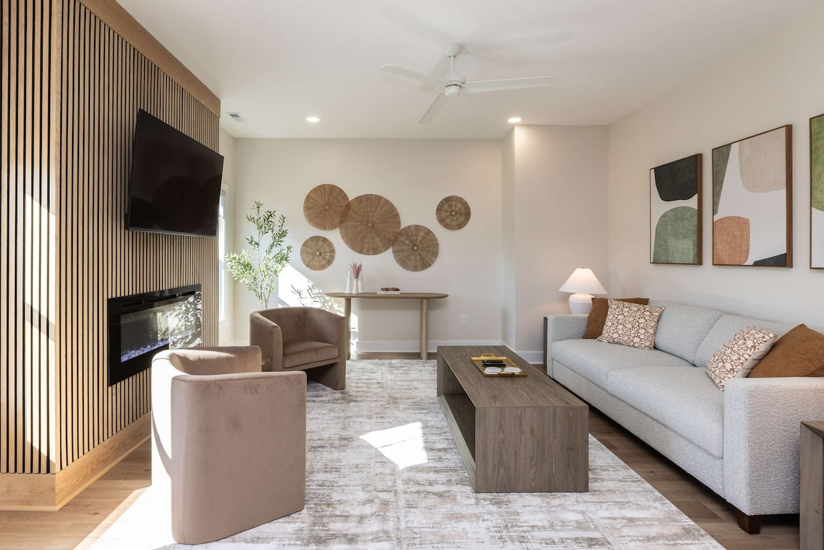 A modern living room features a comfortable sofa and two chairs, illuminated by natural light. Wall art in soft colors is displayed above a sleek coffee table, while a minimalist media unit houses a flat-screen television. A subtle fireplace adds a touch of warmth to the space.