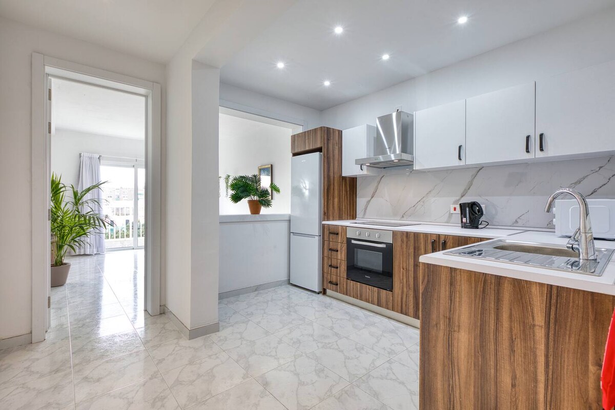 The kitchen features modern cabinetry in warm wood tones, complemented by sleek white countertops. Equipped with essential appliances, including a refrigerator and oven, it offers spacious countertops for meal preparation. Natural light illuminates the area, creating a bright and airy atmosphere.