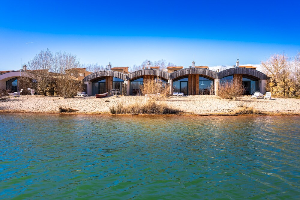 Numerous beach houses are positioned along the shoreline, set against a clear blue sky. The structures feature modern architectural designs, with wooden facades and large windows facing the water. A sparse line of vegetation creates a natural boundary between land and water.