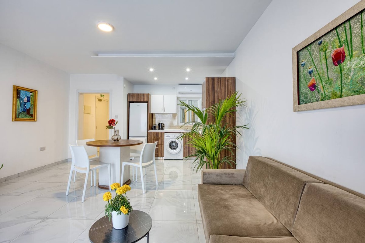 A modern living area is presented with a brown sofa and a small round table surrounded by four white chairs. A decorative plant adds a touch of greenery. The kitchen area, equipped with white cabinetry and appliances, is visible in the background, complemented by light marble flooring.