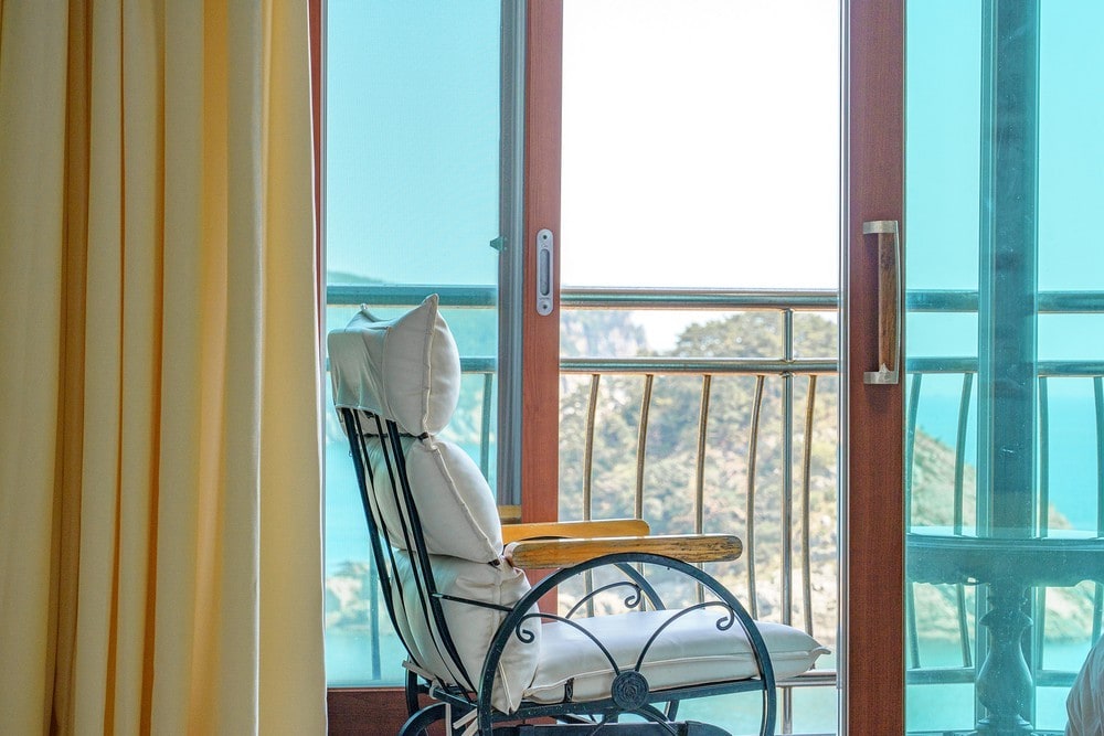 A cozy seating area features a wrought-iron chair with soft cushions, positioned near large glass doors that provide views of the natural landscape. Light-colored curtains frame the doors, allowing sunlight to enter the space.