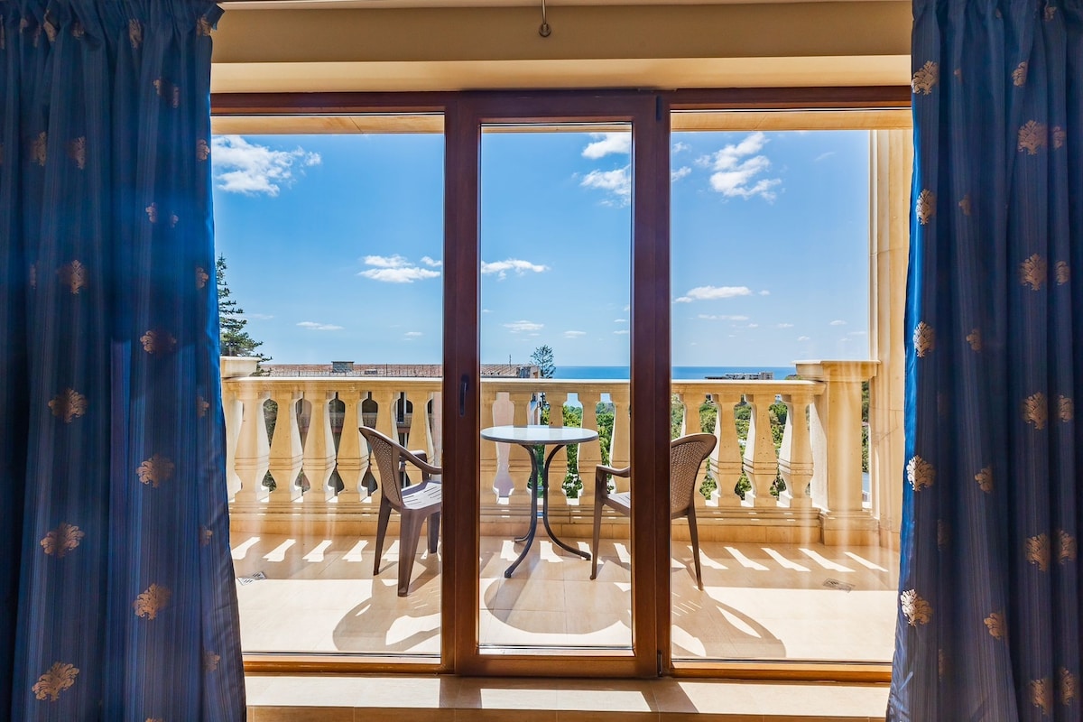 A private balcony is featured, accessed through glass sliding doors. The space includes a small table and two chairs, with an ocean view visible in the distance. The balcony is framed by light blue curtains that softly filter natural light.