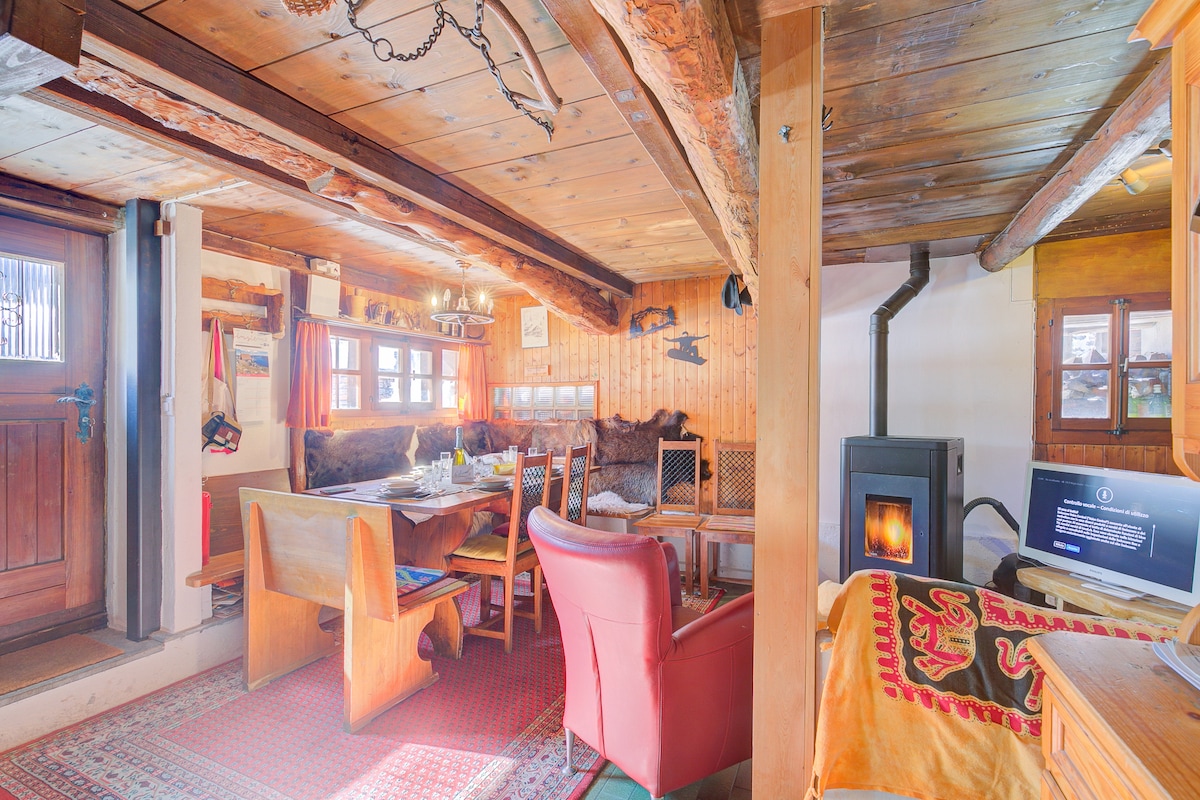 A cozy living room showcases wooden beams and a rustic charm. A dining table for six is set beside a warm pellet stove. Comfortable chairs are arranged around the space, with a digital TV positioned nearby. Natural light filters through the windows, enhancing the inviting atmosphere.