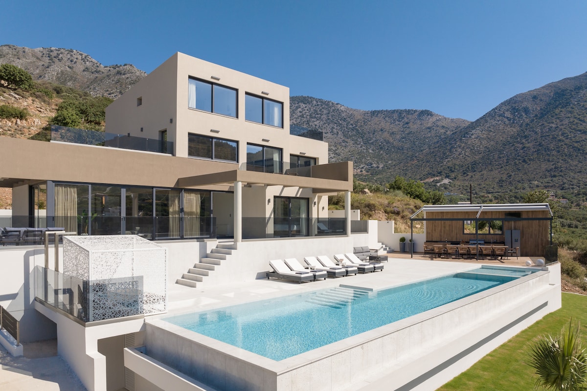 The villa features a stunning infinity pool that mirrors the surrounding mountainous landscape. Expansive terraces are lined with elegant sunbeds, and a shaded outdoor dining area complements the stunning architecture. Large glass windows provide a glimpse into the stylish indoor spaces, enhancing the open-air feel.