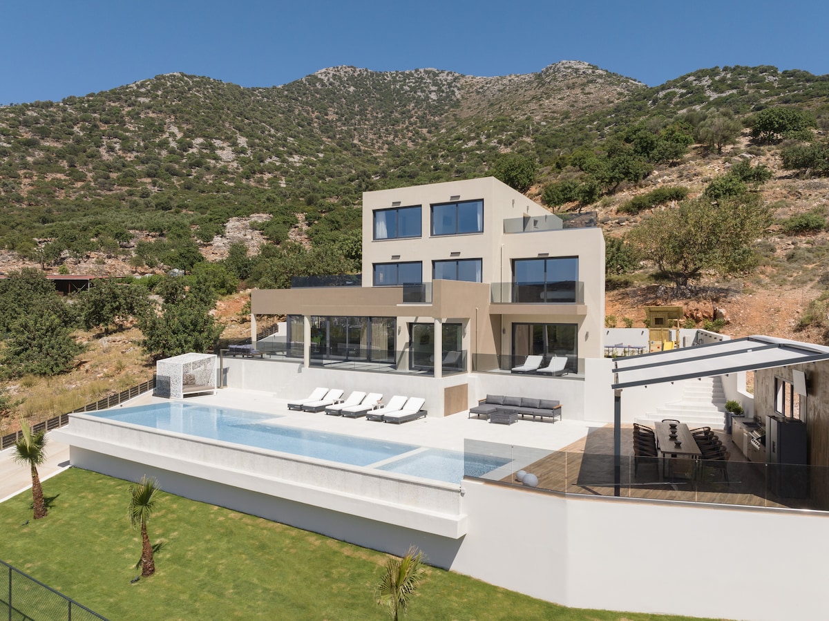 A spacious villa is set against a backdrop of rolling green hills, showcasing a large private infinity pool. Comfortable sun loungers line the pool area, and an outdoor dining space is visible on the terrace, framed by landscaped gardens.