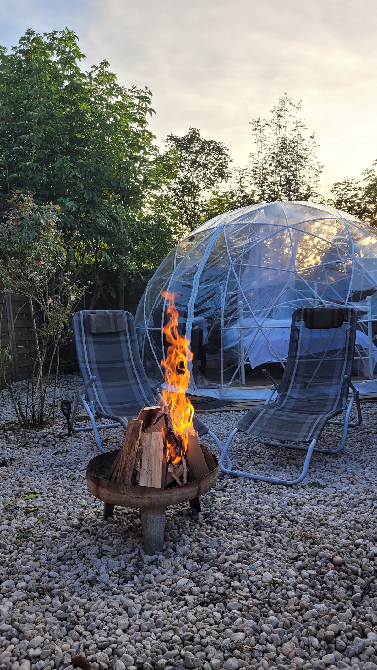 A cozy outdoor area is presented, featuring a fire pit with flames dancing above stacked logs. Two reclining chairs are positioned nearby, with a geodesic bubble structure in the background. Green foliage surrounds the space, creating a serene natural setting.
