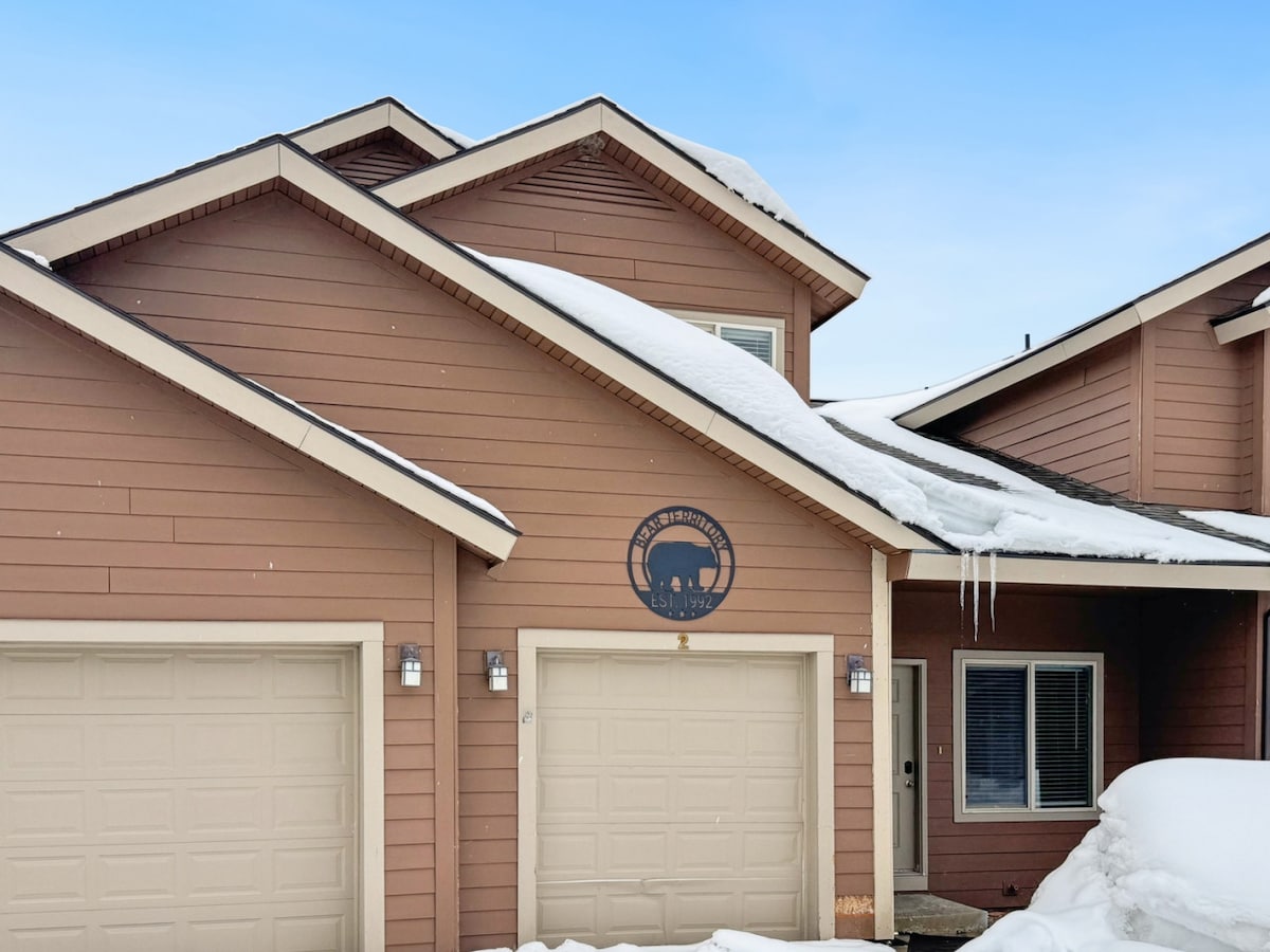 A welcoming townhome exterior features a warm brown facade with a prominent circular emblem. Snow is gently covering the lower areas, while icicles hang from the eaves. The entrance is highlighted by two outdoor lights flanking the door, with a large garage in view.