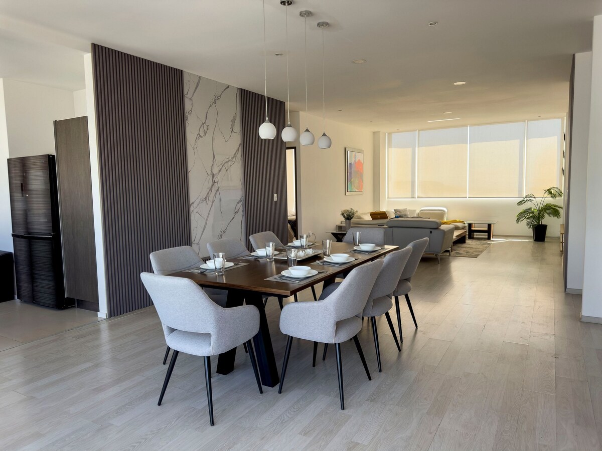 An open dining area features a rectangular wooden table set for six, surrounded by light grey upholstered chairs. Natural light filters through large windows, illuminating the space, which transitions into a cozy living area with a comfortable couch and decorative plants.
