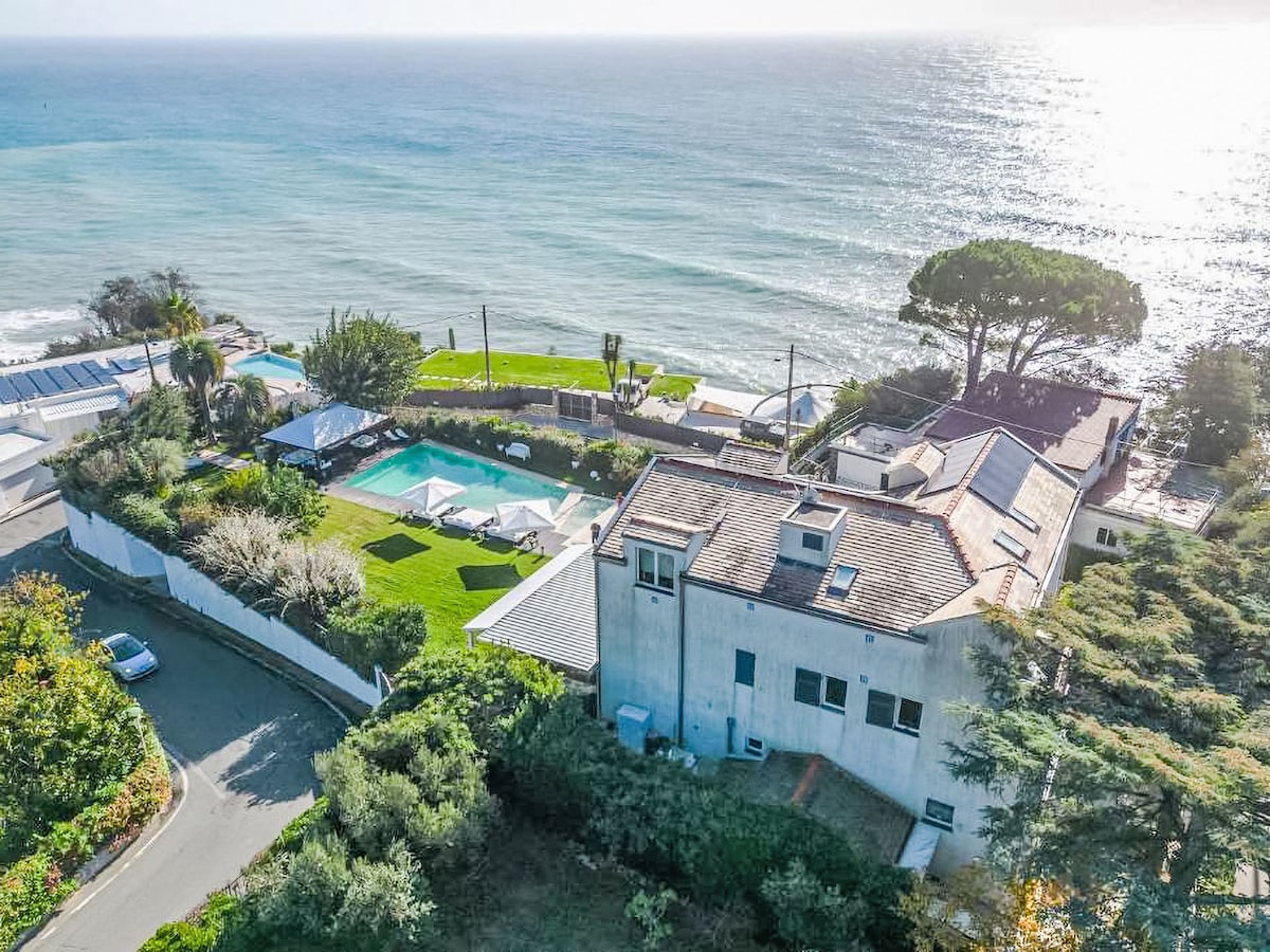 An aerial view of the property showcases the expansive garden and private swimming pool, situated along the coastline. Lush greenery borders the pool area, while the sea sparkles in the background, providing a serene setting for relaxation.