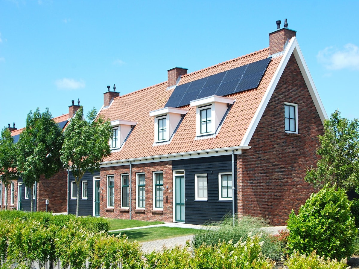 A row of charming houses features a mix of red brick and dark exterior wood. Solar panels are installed on the roof, and green shrubs line the front, accentuating the well-kept lawns. The clear blue sky provides a bright backdrop.