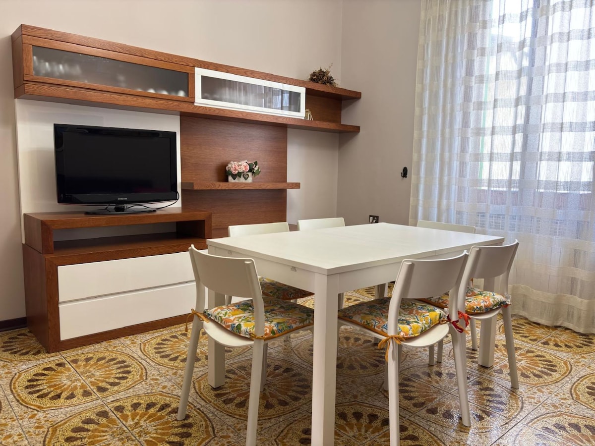 A bright dining area is featured, with a white table surrounded by four chairs. A wooden shelf above the table holds a television and decorative elements. Natural light filters through a large window dressed with sheer curtains, illuminating the patterned floor below.