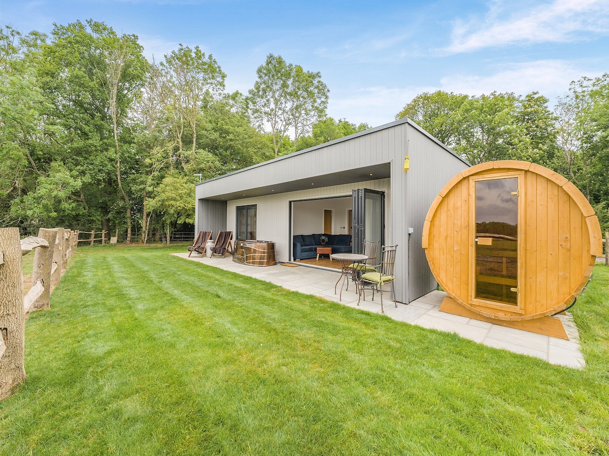 A modern lodge is viewed from the expansive lawn, showcasing floor-to-ceiling glass doors that connect the indoor space with the outdoors. Adjacent, a round wooden sauna sits invitingly, accompanied by outdoor seating and a barbecue area under a clear blue sky and surrounded by lush greenery.