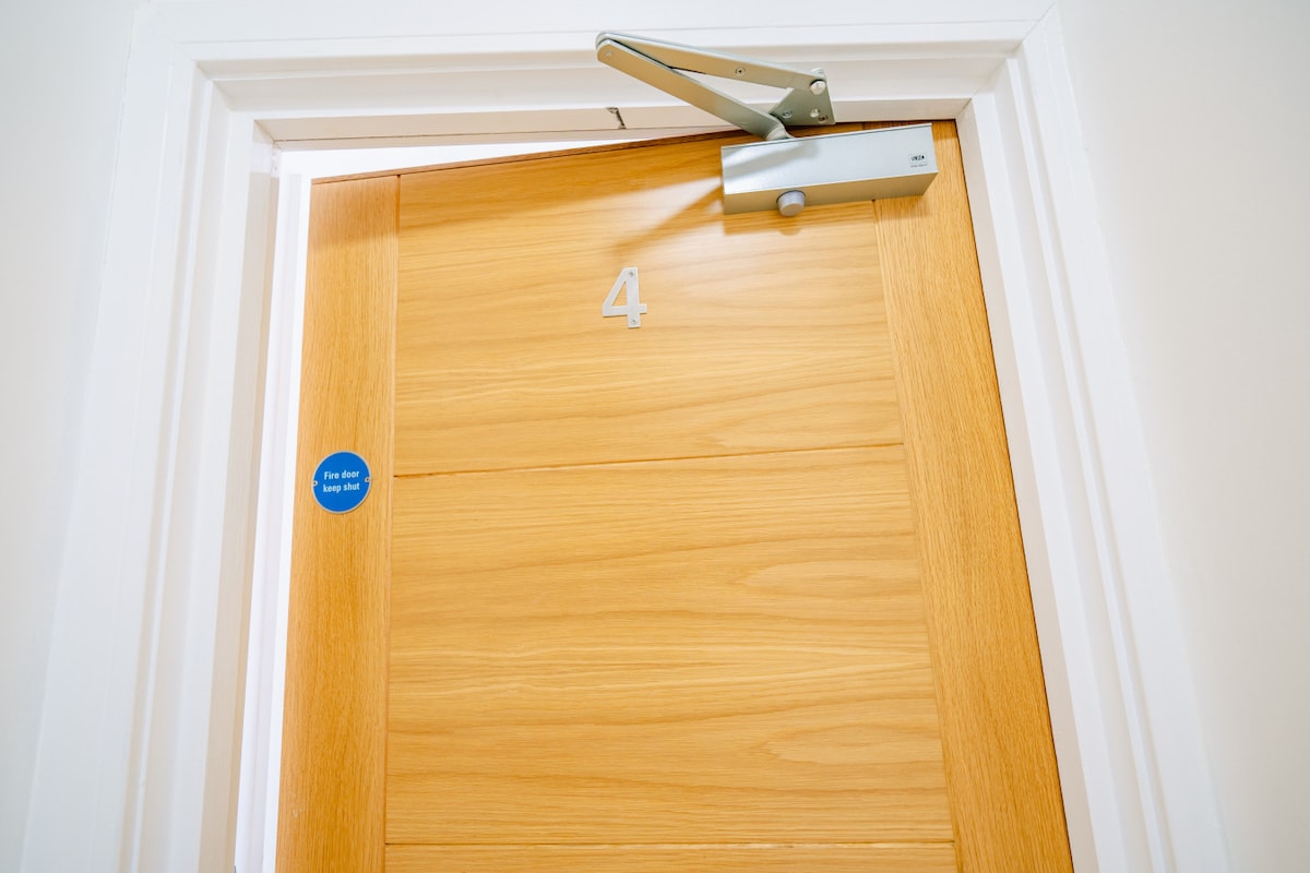 A wooden door is featured, marked with the number '4' in a modern font. A door closer is visible at the top, and a blue circular sticker is placed near the handle, indicating accessibility information.