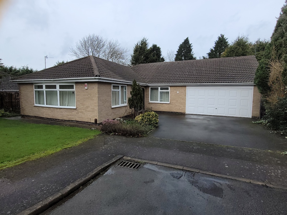 The exterior of a modern bungalow is presented, featuring a light brick facade and large windows. A spacious driveway leads to a double garage. Surrounding greenery includes manicured grass and small bushes, creating a welcoming entrance to the property.