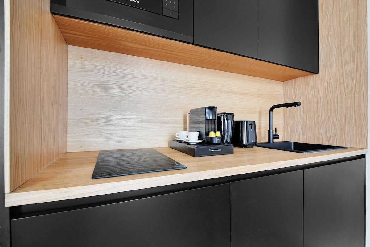 A modern kitchen area features sleek black cabinetry and a wooden countertop. Kitchen appliances include a coffee machine, kettle, and a few ceramic cups, neatly arranged on the counter, with a black sink positioned alongside for convenience.