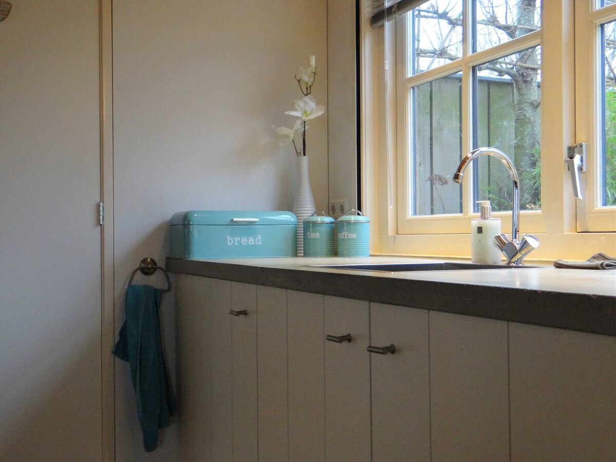 A kitchen area features a light-colored countertop with a sleek sink and modern faucet. Two decorative storage containers in soothing teal are placed alongside a simple arrangement of flowers. Natural light filters through the nearby windows, illuminating the space.