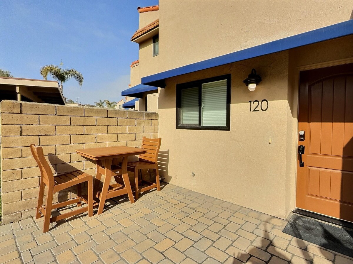 A private patio features a small wooden table accompanied by two chairs, set against a textured brick wall. The sunlit area is sheltered by a blue awning, while a warm-toned door is partially visible, displaying the unit number 120.
