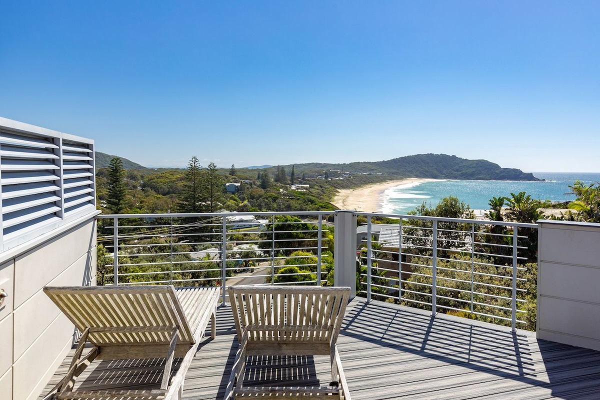 An inviting rooftop deck features two reclining lounge chairs positioned for optimal views of the surrounding landscape. The sandy beach curves along the coastline, framed by rolling hills under clear blue skies, offering a serene escape for relaxation.