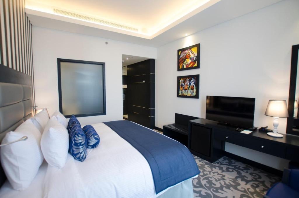 A modern hotel room features a spacious bed with crisp white linens and a blue decorative blanket. Colorful artwork adorns the walls, and a sleek television is positioned on a dark console. An illuminated lamp adds a touch of warmth to the ambiance.