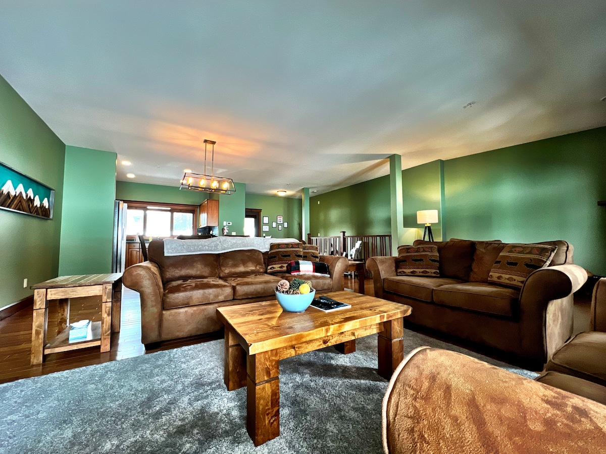 The open living area features comfortable brown sofas arranged around a wooden coffee table, which is highlighted by a decorative bowl. Soft green walls provide a calming backdrop, while natural light fills the space, enhancing the inviting ambiance.