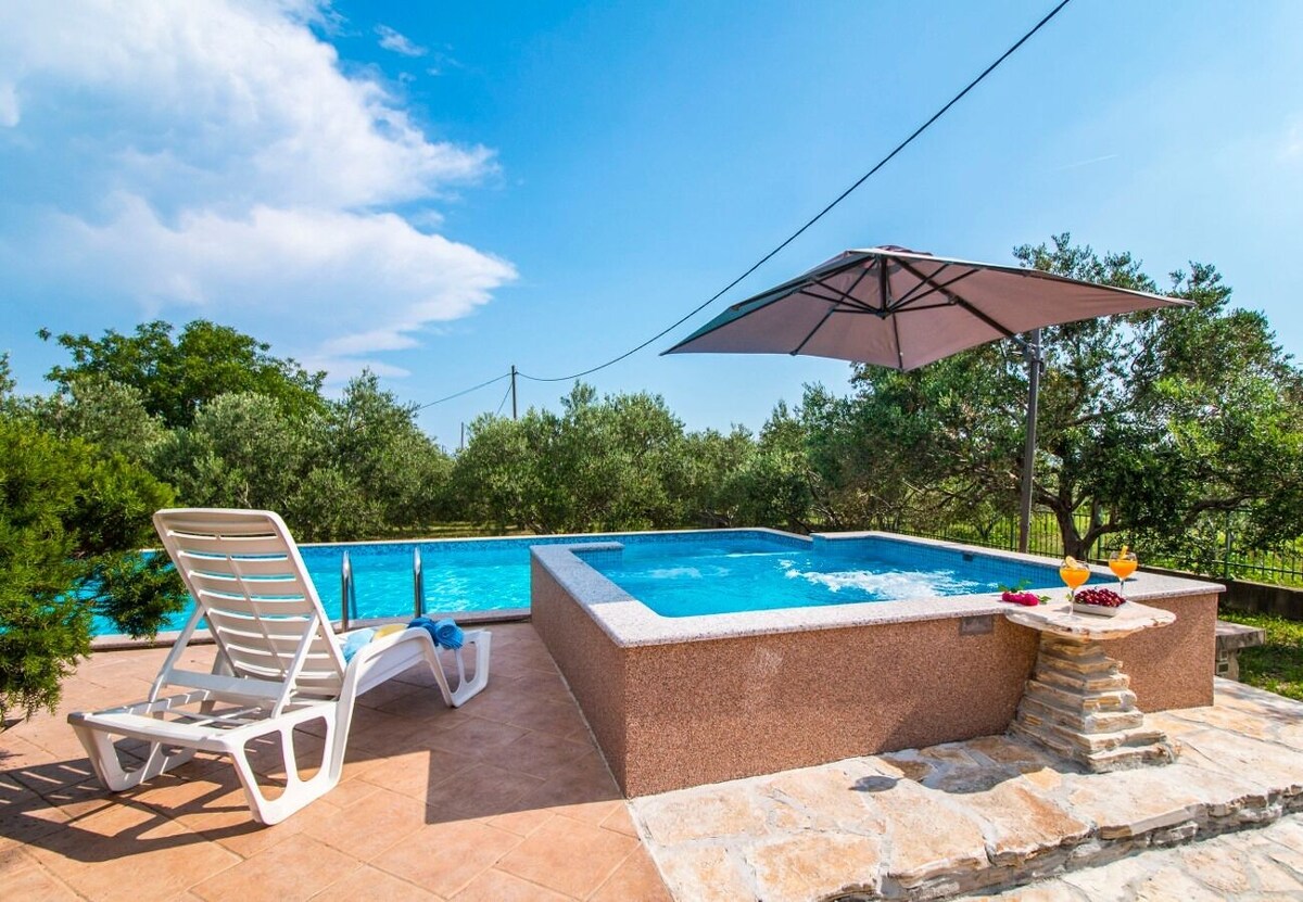 A bright outdoor space features a refreshing pool accompanied by a smaller jacuzzi. A large umbrella provides shade to a nearby lounge chair, while a snack spread awaits on a stone table. Lush greenery surrounds the area, contributing to a serene atmosphere.