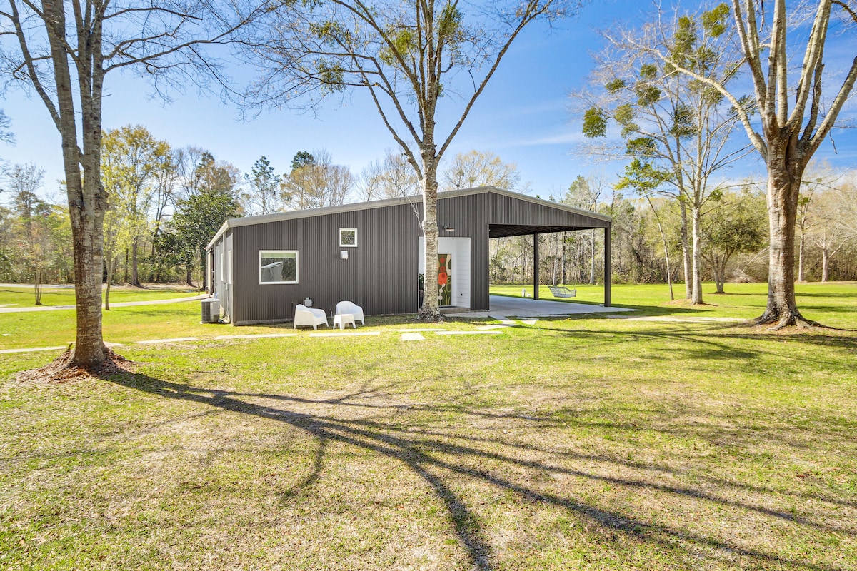 A spacious yard surrounds a modern barndominium, featuring a covered patio and two chairs placed on the green grass. Trees provide shade, and the exterior showcases a simple aesthetic with large windows and a clear blue sky above.