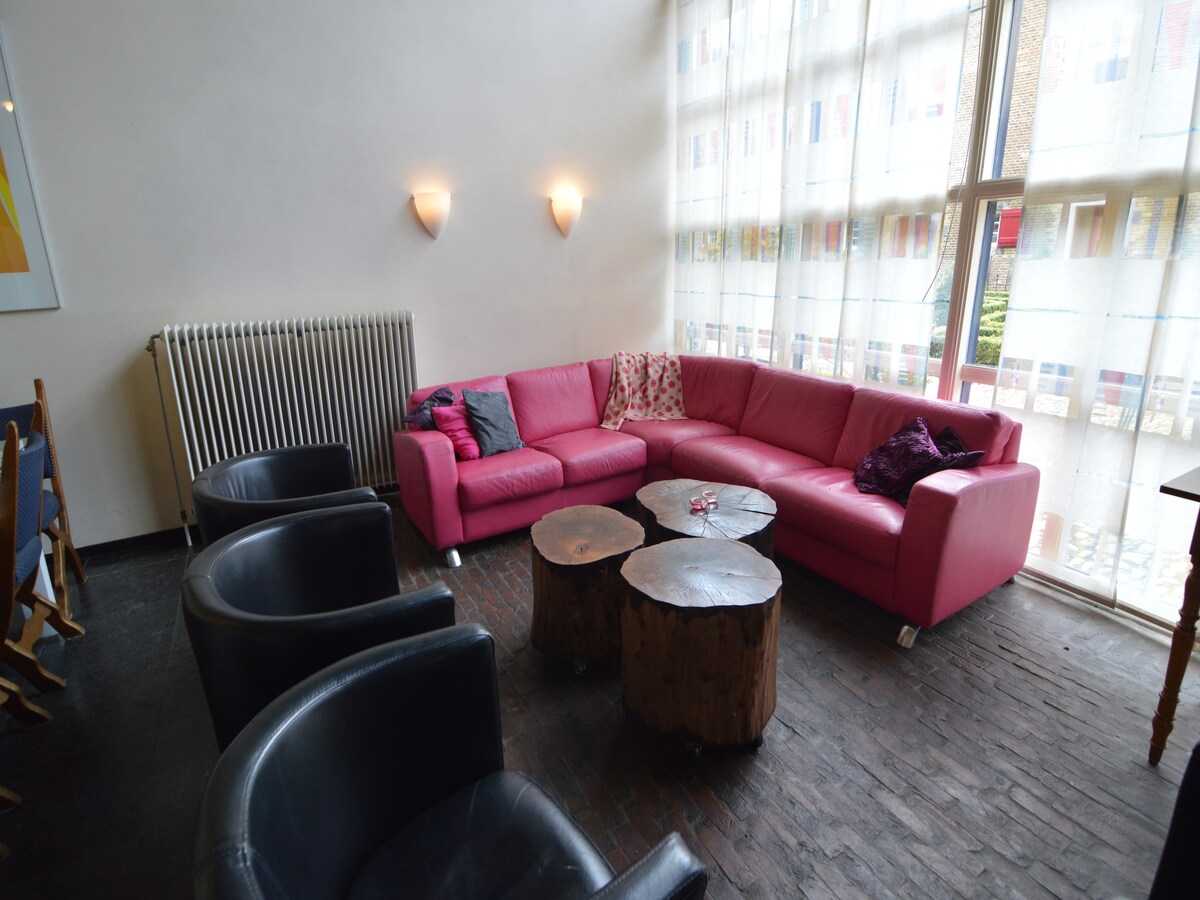 A bright living area is showcased, featuring a large pink sectional sofa with decorative pillows. Two wooden coffee tables contribute to the cozy setup, while black armchairs provide additional seating options. Natural light filters through large windows, creating an open and welcoming atmosphere.