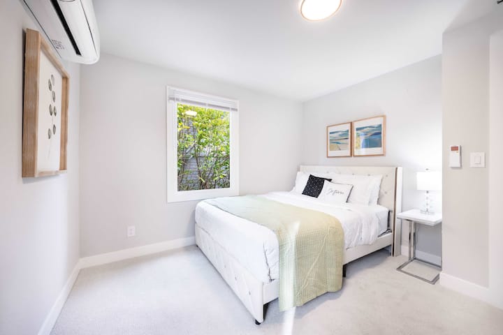 Уютна спалня с 2 модерни картини, плюшено легло с меко спално бельо и стилни нощни шкафчета. Топлата светлина и прозорецът създават светла и привлекателна атмосфера.