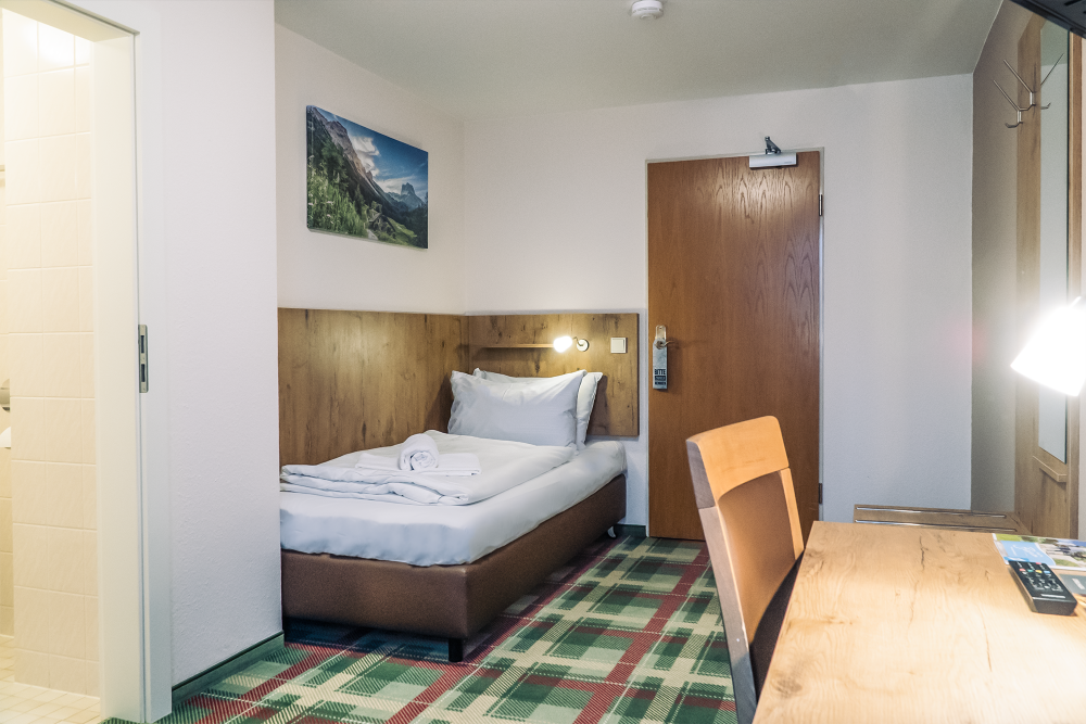 A single room features a spacious bed with fresh linens, positioned against a wooden headboard. A desk with a lamp is included, along with a wooden chair. A bathroom door is visible in the background, and the patterned carpet adds a touch of comfort.