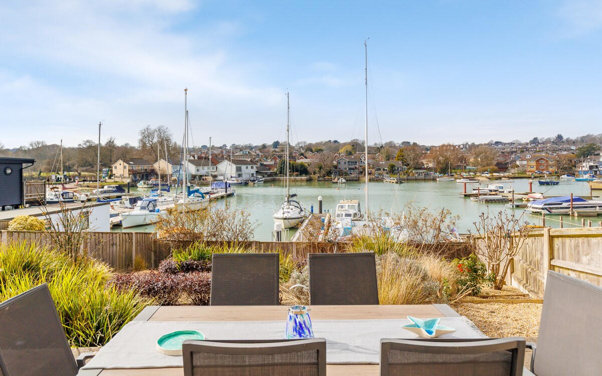 An outdoor dining area features a spacious table with chairs, set against a backdrop of a marina. Various boats are visible on the calm water, framed by greenery and a clear blue sky. The scene conveys a peaceful waterfront atmosphere.