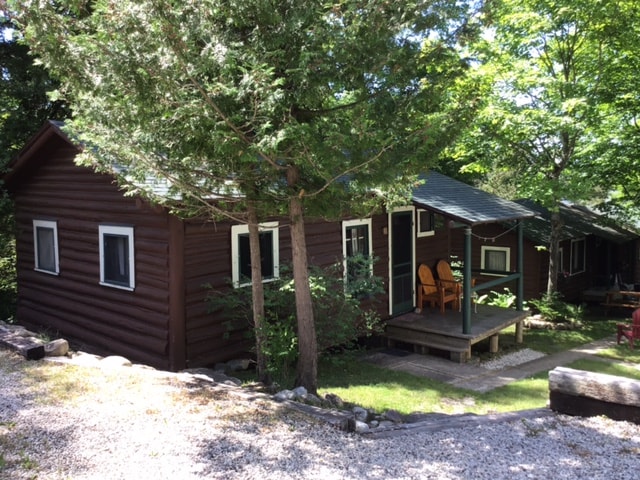 The log cabin is framed by lush greenery, featuring a welcoming porch with two chairs facing the entry. Large windows allow natural light to illuminate the cabin's exterior, while a stone path leads through the yard, highlighting the cabin's rustic charm.