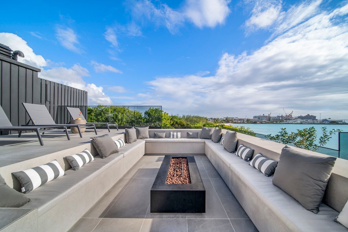 A rooftop lounge area is designed with a modern fire pit at its center, surrounded by built-in seating adorned with plush gray cushions and striped accents. Lounge chairs are positioned for sunbathing, with views of the ocean and coastal landscape in the background.