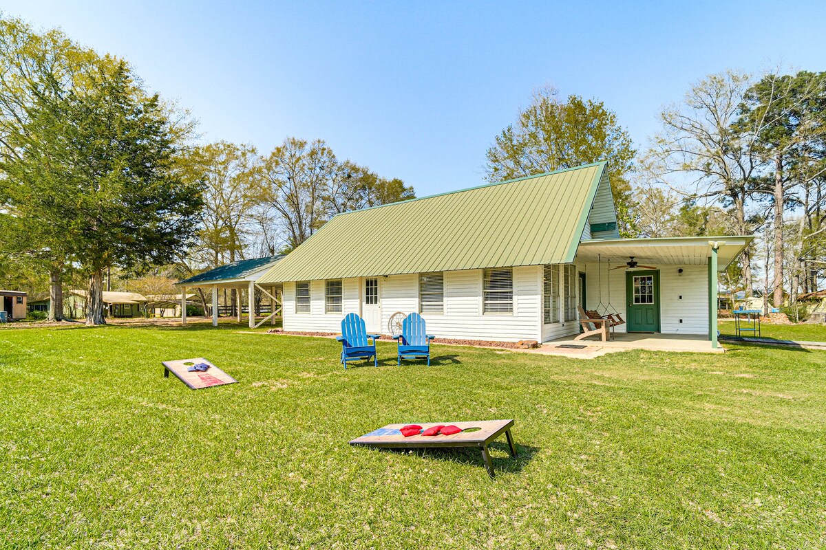 A large, well-maintained cabin is shown with a green metal roof. Two blue adirondack chairs are positioned on the green lawn, which features a cornhole setup. Trees surround the property, and a spacious porch is visible, enhancing outdoor living opportunities.