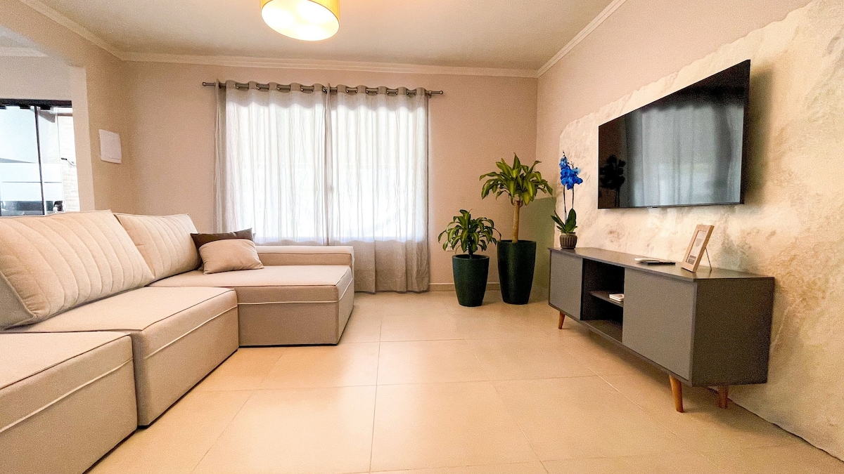 A welcoming living area features a spacious sectional sofa, complemented by a modern TV mounted on a textured wall. Natural light enters through sheer curtains, and indoor plants add a refreshing touch to the room’s neutral tones.