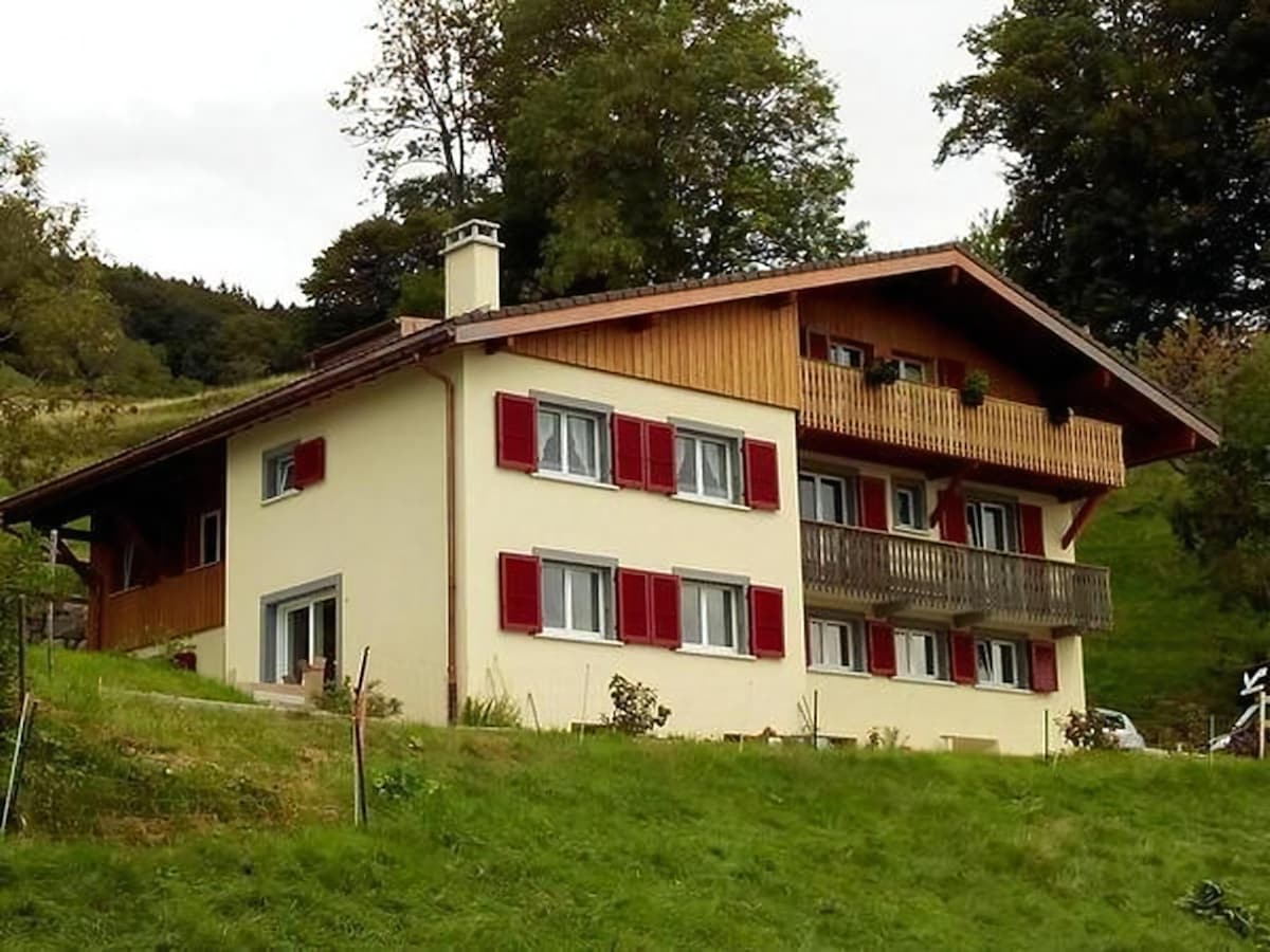 A two-story house features a mix of wooden and stucco exteriors, with red shutters enhancing the window areas. A balcony spans the upper level, surrounded by greenery. The property is set within a lush, grassy landscape, providing a tranquil rural setting.