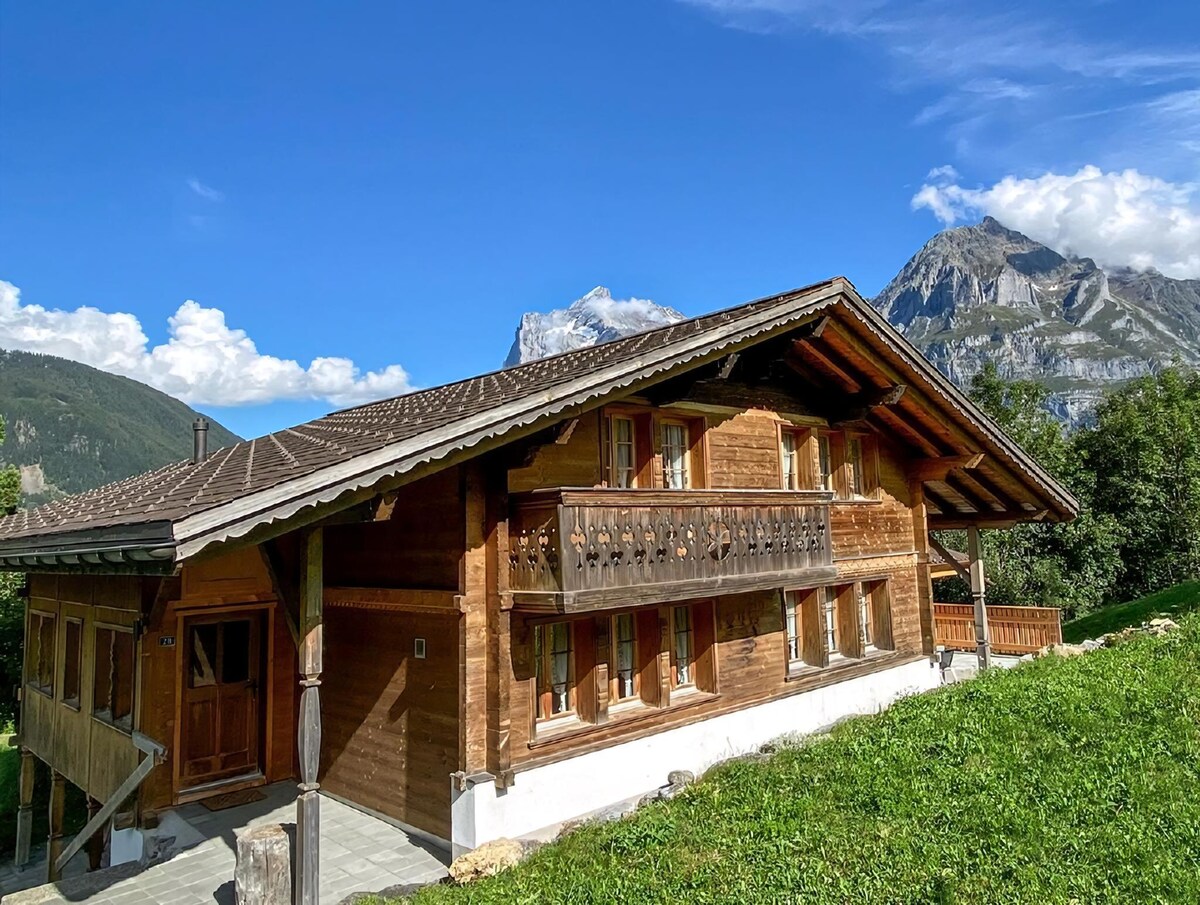 The charming chalet is set against a backdrop of majestic mountains under a clear blue sky. Traditional wooden architecture is featured, along with a balcony offering views of the surrounding landscape. Lush green grass lines the front of the property, enhancing the natural setting.