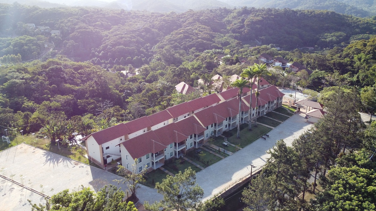 An aerial view of the apartment complex reveals a row of two-story buildings with red roofs nestled among greenery. Surrounding trees and landscaped areas enhance the natural setting, while open pathways lead through the property, providing access to each unit.