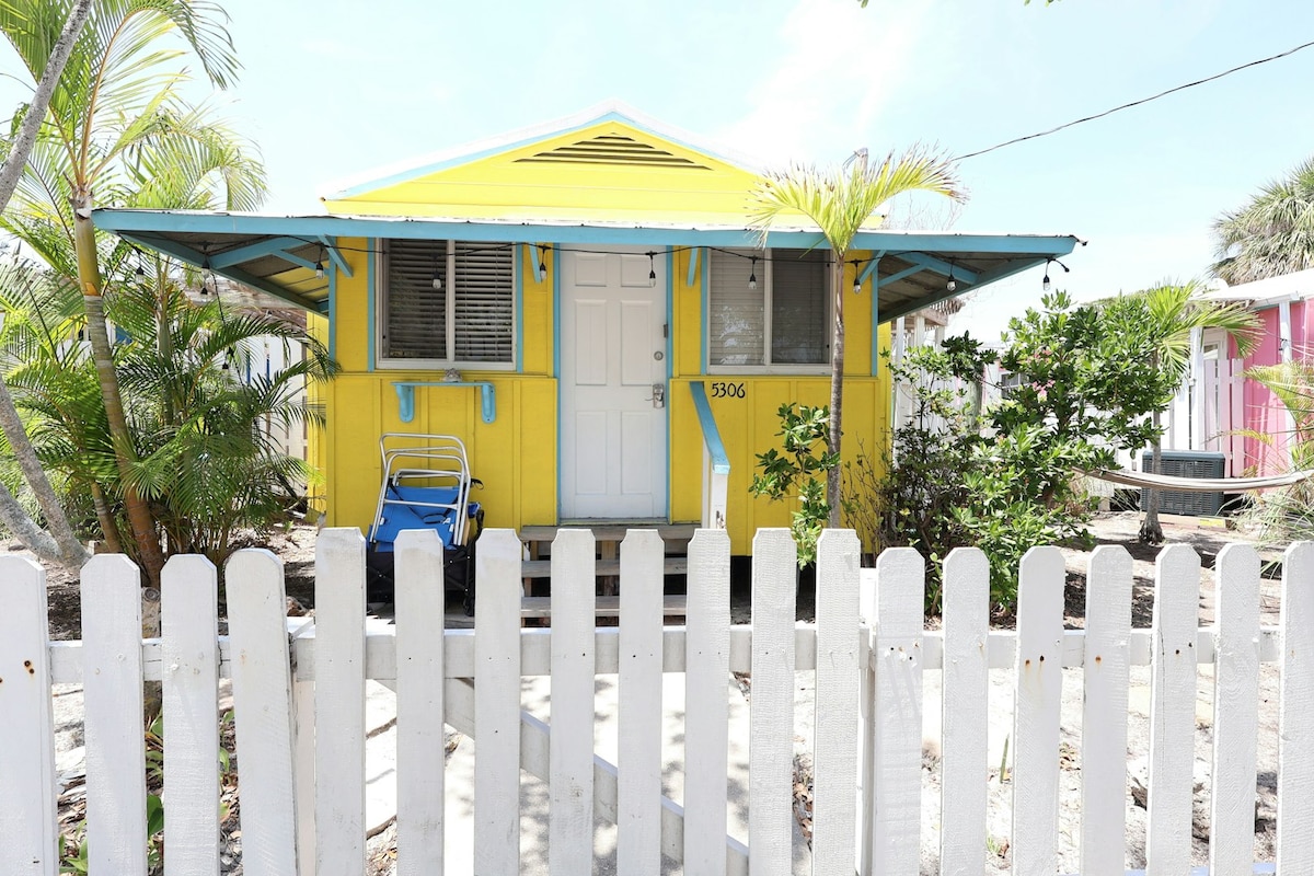 A charming yellow cottage is framed by a white picket fence, with tropical plants and palm trees surrounding the entrance. The front door is complemented by blue accents, while a blue folding chair is positioned nearby, enhancing the inviting exterior.