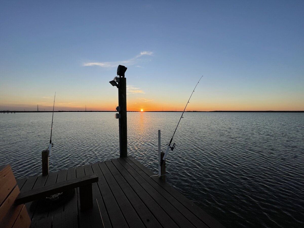 A serene view from a fishing dock at sunset, with two fishing rods positioned to the sides. The calm water reflects shades of orange and blue from the sky, while wooden planks create a sturdy base. A light fixture is visible, enhancing the area for evening use.