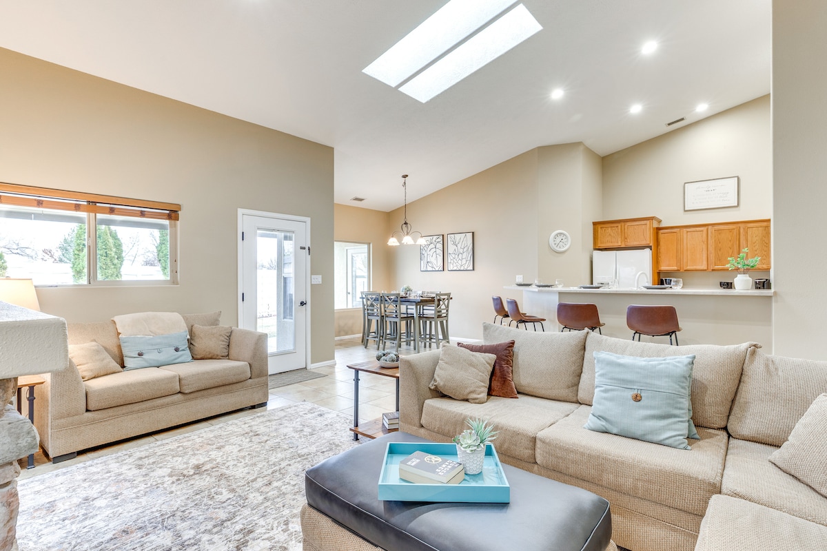 A spacious living area is showcased with natural light filtering through skylights and large windows. Comfortable seating is arranged around a central coffee table, while a dining area with high-top seating is visible in the background, complemented by wooden cabinetry in the kitchen.