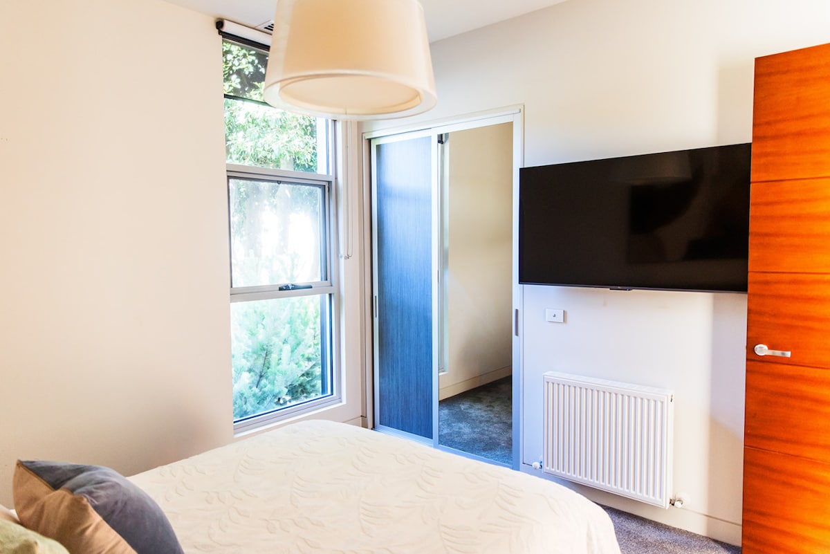 Štýlová spálňa s manželskou posteľou, televízorom a panelovým ohrievačom.